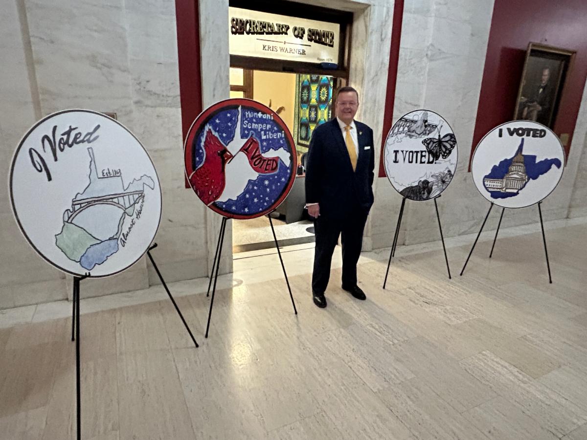 Kris Warner standing in front of the I Voted sticker finalists displayed at the Secretary of States office