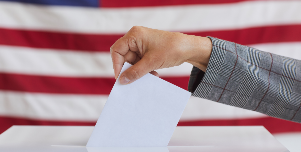 Hand placing a ballot into a ballot box with an American flag backdrop